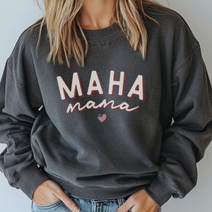 May include: Pepper-colored sweatshirt featuring the text 'MAHA mama' in a stylized font with a small American flag heart graphic. The sweatshirt is a crewneck style with long sleeves and a relaxed fit.  It's made of a soft, comfortable fabric suitable for casual wear.