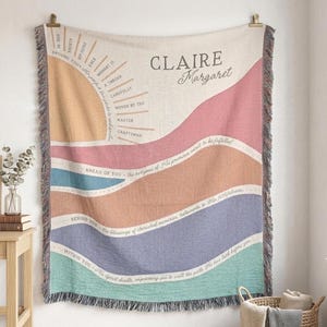 May include: A woven throw blanket with a colorful landscape design. The blanket features a sun graphic and text that reads "Claire Margaret." The design includes shades of peach, pink, blue, and green, with a fringed edge.