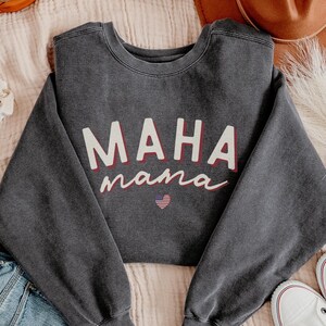 May include: A dark gray sweatshirt with the text "MAHA mama" in white with a red outline. A small American flag is inside a red heart.