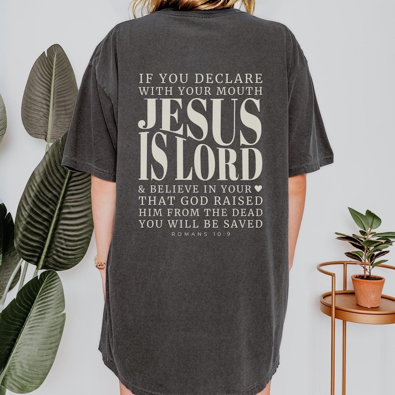 以下が含まれることがあります： 「JESUS IS LORD」の文字が白の太字でプリントされた、濃いグレーのTシャツ。背中には「IF YOU DECLARE WITH YOUR MOUTH...YOU WILL BE SAVED」と「ROMANS 10:9」の文字もプリントされています。