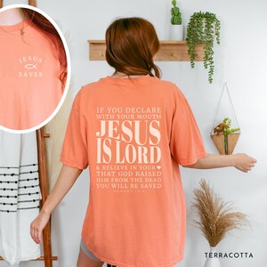 以下が含まれることがあります： テラコッタ色のTシャツ。前面には「Jesus Saves」の文字と魚のシンボルがプリントされています。背面には「Jesus Is Lord」の文字と聖書の言葉が書かれています。快適なオーバーサイズフィットです。