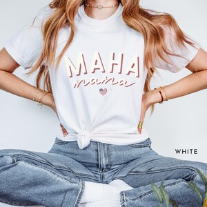 May include: White t-shirt with the text "MAHA" in pink and the text "inspired" in a cursive font with a small American flag heart.
