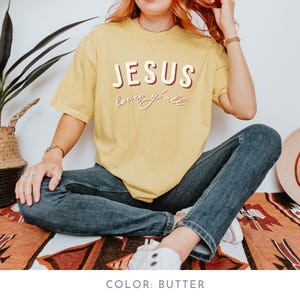 May include: A butter-yellow t-shirt featuring the text "JESUS loves y'all" in a retro font. The shirt is short-sleeved and casual. The model is wearing blue jeans and white trainers. The shirt is made of a soft, comfortable material.