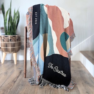 May include: Woven throw blanket with an abstract geometric design in shades of blue, teal, peach, and black. The blanket has fringed edges and features the text "EST 2026" and "The Castros" in white.