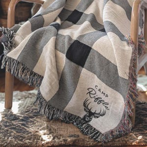 May include: A woven throw blanket with a black, white, and gray checkered pattern. The blanket has a fringed edge and the words "CAMP Riffle" with a deer head graphic are displayed on the corner. The blanket is draped over a wooden chair.
