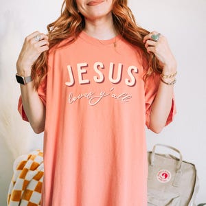 May include: Coral-coloured t-shirt featuring the text "JESUS loves y'all" in a layered, serif font. The shirt has a relaxed fit and short sleeves. The image shows the shirt being worn.