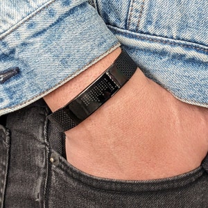 May include: Black mesh bracelet with a rectangular plate engraved with the words "MY TRIBE" and several names. The bracelet is worn on a wrist, with a denim jacket and black jeans in the background.