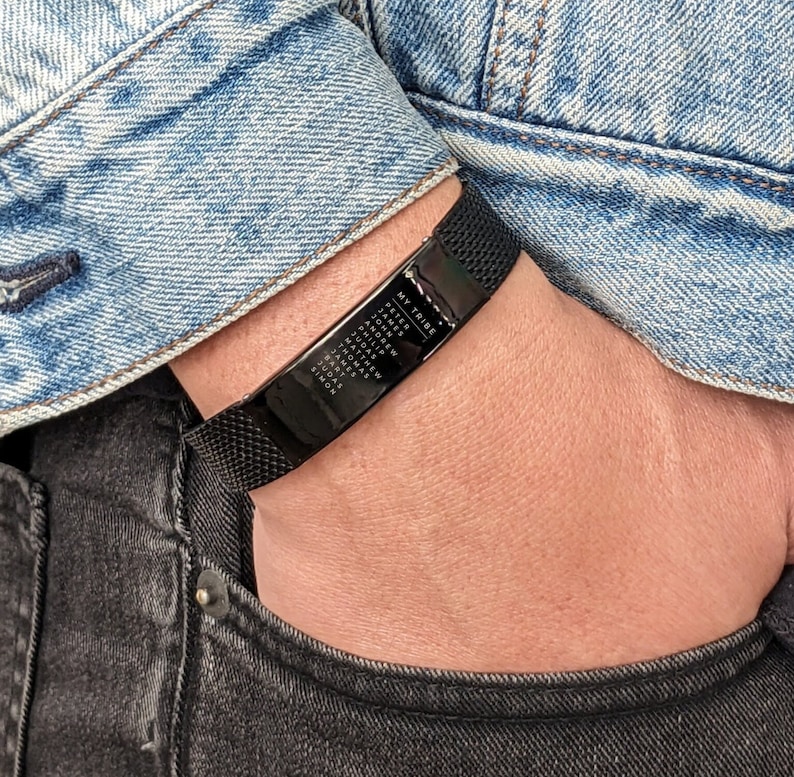 May include: Black mesh bracelet with a rectangular plate engraved with the words "MY TRIBE" and several names. The bracelet is worn on a wrist, with a denim jacket and black jeans in the background.