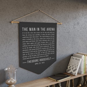May include: Black and white canvas wall tapestry with a quote by Theodore Roosevelt, "The Man in the Arena", from April 23, 1910. The quote is about the importance of action and striving for greatness.