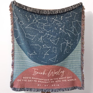 May include: A woven throw blanket with a celestial design. The navy blue blanket features white constellations and the text "Isaiah Wesley" with a date. The blanket has a fringed edge and a half-circle design in shades of blue and red.