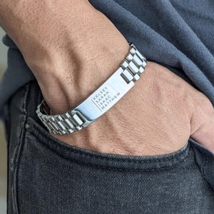 May include: A silver-toned stainless steel bracelet with a rectangular nameplate. The nameplate is engraved with the names "KELSEY", "SARAH", "ISAAC", and "MATTHEW". The bracelet is worn on a wrist, with the hand in a pocket.