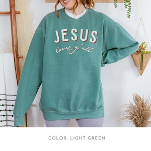 May include: Light green sweatshirt with the text "JESUS loves y'all" in white and red lettering. The sweatshirt has a crew neck and long sleeves. The colour is light green.