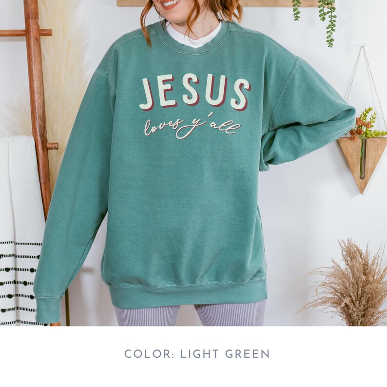 May include: Light green sweatshirt with the text "JESUS loves y'all" in white and red lettering. The sweatshirt has a crew neck and long sleeves. The colour is light green.