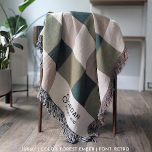 Puede incluir: Una manta tejida con un patr&oacute;n de diamantes en tonos verdes, beige y crema, sobre una silla de madera. La manta mide 127x152 cm y tiene un ribete de flecos. El nombre "Jordan" y el texto "Isa&iacute;as 41:10" est&aacute;n impresos en la manta.