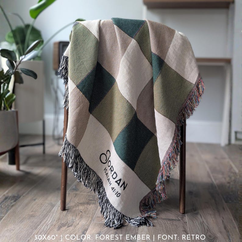 Puede incluir: Una manta tejida con un patr&oacute;n de diamantes en tonos verdes, beige y crema, sobre una silla de madera. La manta mide 127x152 cm y tiene un ribete de flecos. El nombre "Jordan" y el texto "Isa&iacute;as 41:10" est&aacute;n impresos en la manta.
