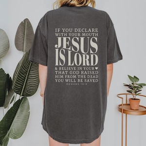 以下が含まれることがあります： クリーム色の「JESUS IS LORD」の文字が入った濃いグレーのTシャツ。テキストには聖書のローマ人への手紙10:9が含まれています。ゆったりとしたフィット感のシャツです。