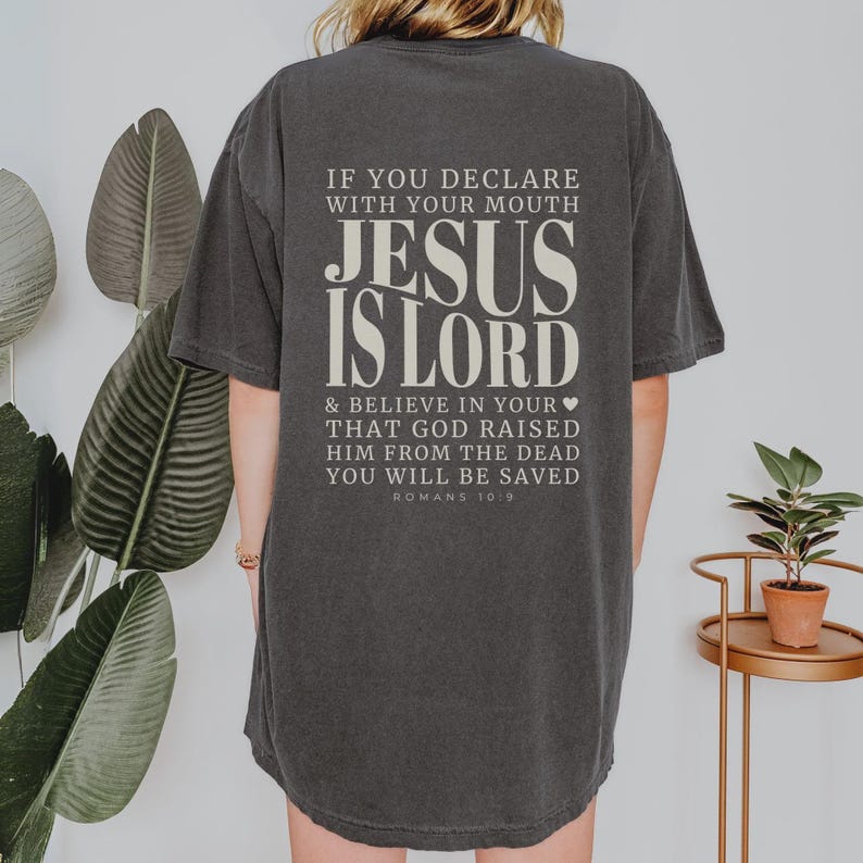以下が含まれることがあります： クリーム色の「JESUS IS LORD」の文字が入った濃いグレーのTシャツ。テキストには聖書のローマ人への手紙10:9が含まれています。ゆったりとしたフィット感のシャツです。