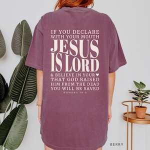 以下が含まれることがあります： 大胆で様式化されたフォントで「JESUS IS LORD」というテキストがプリントされたベリー色のTシャツ。Tシャツの背面には、ローマ人への手紙10:9からの宗教的な引用があります。このシャツは快適なオーバーサイズフィットです。