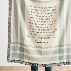May include: A woven throw blanket with a light green and white plaid pattern. The blanket features the text of Psalm 23:1-4 in black script. The edges of the blanket have a fringed trim.
