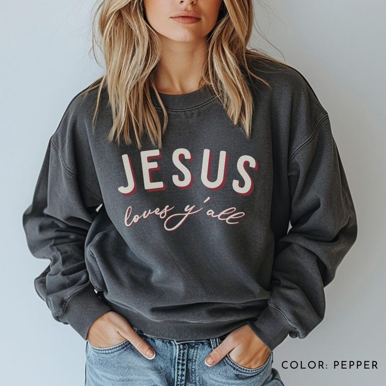 May include: A dark grey sweatshirt with the text "JESUS loves y'all" printed on the front in white and red lettering. The sweatshirt is a crew neck style and has long sleeves. The colour is pepper.