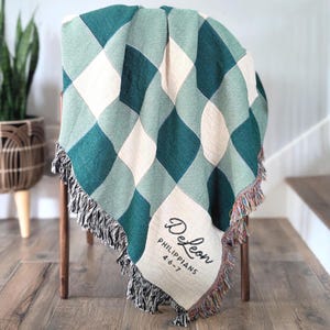 May include: A woven throw blanket draped over a wooden chair. The blanket features a diamond pattern in shades of green and cream, with a fringed edge. The text "DeLeon" and "Philippians 4:6-7" are embroidered on the corner.