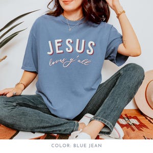 May include: A blue jean coloured t-shirt with the words "JESUS loves y'all" in white and red lettering. The shirt is paired with blue jeans and white trainers. The overall theme is religious and casual.