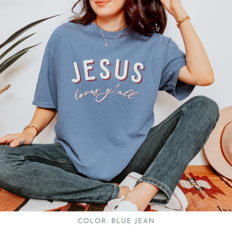 May include: A blue jean coloured t-shirt with the words "JESUS loves y'all" in white and red lettering. The shirt is paired with blue jeans and white trainers. The overall theme is religious and casual.