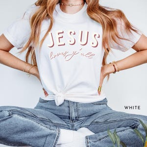 May include: White t-shirt featuring the text "JESUS loves y'all" in burgundy and cursive lettering. The shirt has a crew neck and short sleeves. The model is wearing blue jeans.