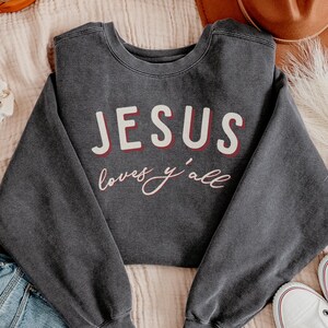 May include: Dark grey crew neck sweatshirt featuring the text "JESUS loves y'all" in white and red. The sweatshirt is made of a soft material. The text is centered on the front of the garment, with the words in a bold font.