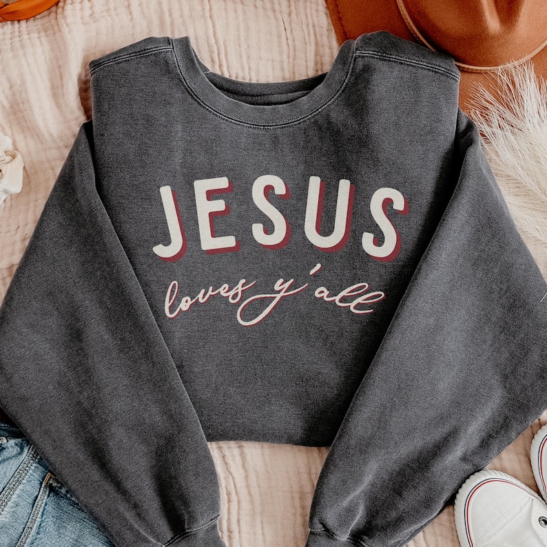 May include: Dark grey crew neck sweatshirt featuring the text "JESUS loves y'all" in white and red. The sweatshirt is made of a soft material. The text is centered on the front of the garment, with the words in a bold font.