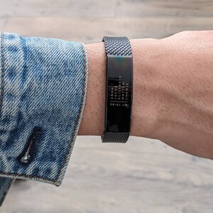May include: A black metal mesh bracelet with a rectangular plate. The plate has the words "ANNA JACOB ELIZABETH MATTHEW CHEWIE" and "MY TRIBE" in white text. The bracelet is worn on a wrist, with a denim jacket sleeve in the background.