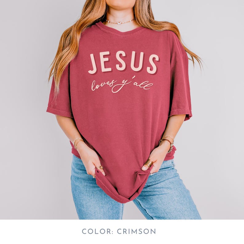 May include: Crimson t-shirt featuring the text "JESUS loves y'all" in a layered, off-white font. The shirt has a relaxed fit and a v-neck. The model is wearing blue jeans.