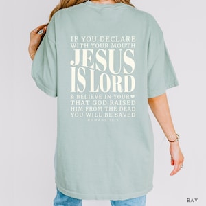 以下が含まれることがあります： 「JESUS IS LORD」の文字が大きくオフホワイトでプリントされた、薄い緑色のTシャツ。背中には「IF YOU DECLARE WITH YOUR MOUTH...YOU WILL BE SAVED」と「ROMANS 10:9」の文字もプリントされています。