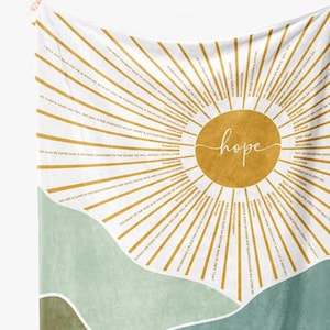 May include: White and gold blanket with the word "hope" in cursive within a gold sunburst design. The blanket features inspirational quotes and a landscape design in shades of green and brown.