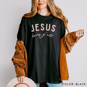 May include: Black crew neck t-shirt featuring the text "JESUS loves y'all" in white and red. The shirt is worn by a person. The t-shirt is black in colour.