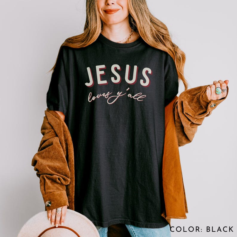 May include: Black crew neck t-shirt featuring the text "JESUS loves y'all" in white and red. The shirt is worn by a person. The t-shirt is black in colour.