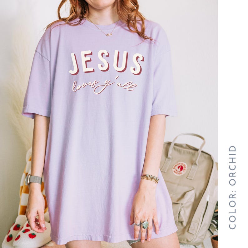 May include: Orchid-coloured t-shirt with the text "JESUS loves y'all" in a two-tone design. The shirt is a loose fit and has short sleeves. The colour is described as orchid.