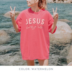 May include: Watermelon-coloured t-shirt with the text "JESUS loves y'all" in a layered design. The shirt is a solid colour and has a relaxed fit. The person in the image is giving a peace sign.