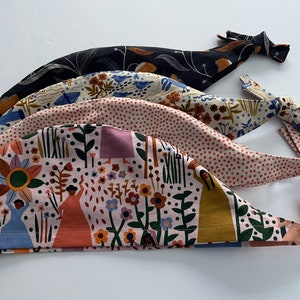 May include: Four colourful fabric scrub hats with different patterns, including a floral design with women and a polka dot pattern. The hats are tied at the back with long ties.