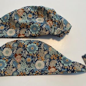 May include: Two blue floral patterned scrub caps. One is a bouffant style, and the other is a tie-back style. The fabric features a repeating pattern of flowers, faces, and foliage in shades of blue, white, and brown.