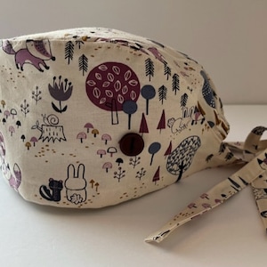 May include: A beige scrub hat with a woodland print featuring foxes, rabbits, squirrels, mushrooms, and trees. The hat has a button on the top and ties at the back.