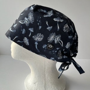 May include: A black scrub hat with a white mushroom print. The hat has a button on the top and ties at the back.