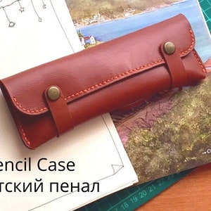 Peut inclure: DIY Étui à crayons minimaliste, un étui à crayons en cuir marron avec une sangle et une fermeture à bouton. L'étui est posé sur un carnet avec un dessin d'une maison et d'une étendue d'eau.