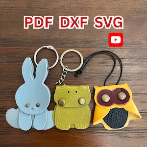 Può includere: Tre portachiavi in pelle: un coniglietto azzurro, un ippopotamo giallo-verde e un gufo giallo con dettagli marroni. Ogni portachiavi ha un anello di metallo. Il testo "PDF DXF SVG" è in alto.
