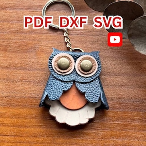 Könnte beinhalten: Ein eulenformiger Schlüsselanhänger aus strukturiertem dunkelblauem Leder mit hellbraunem Bauch und beigefarbener, gewellter Basis. Die Augen bestehen aus beigen Lederkreisen mit messingfarbenen Metallnieten. Der Text PDF DXF SVG ist oben sichtbar.