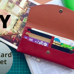 May include: DIY Long Card Wallet, a red leather wallet with multiple card slots, open to show the interior and several cards inside. The wallet is on a green cutting mat with a magazine and a green book in the background.