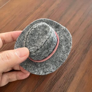 Op de afbeelding: Een kleine, ronde hoed van grijze vilt met een platte kroon en een brede rand. De hoed is versierd met een dunne, lichtroze leren band die rond de basis van de kroon is gewikkeld en vastgezet met zichtbare stiksels langs de bovenrand.