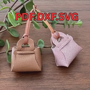 May include: Two miniature leather handbag charms, one tan and one pink, each with a handle and a leather strap. The text "PDF.DXF.SVG" is displayed in red above the charms. Green leaves and brown branches are in the background.