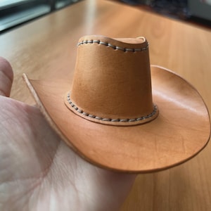 以下が含まれることがあります： 明るい茶色の革で作られたミニチュアのカウボーイハット。丸みを帯びたクラウンと広いブリムが特徴で、縁には装飾的なステッチが施されています。この帽子は小さく、ディスプレイやノベルティアイテムに適しています。