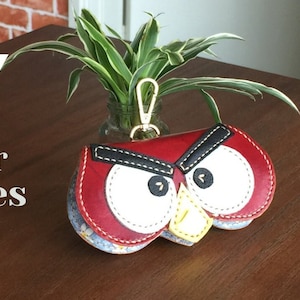 May include: A red leather Angry Birds-themed keychain with white eyes, a black eyebrow, and a yellow beak. The keychain is attached to a gold metal ring.